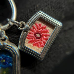 May include: Close-up of a silver charm with a rectangular frame. Inside the frame is a small, embroidered red flower on a beige background. The charm is attached to a silver ring.
