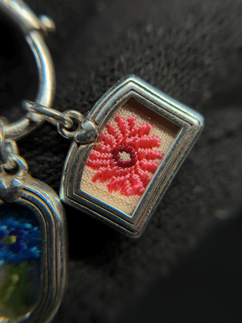 May include: Close-up of a silver charm with a rectangular frame. Inside the frame is a small, embroidered red flower on a beige background. The charm is attached to a silver ring.