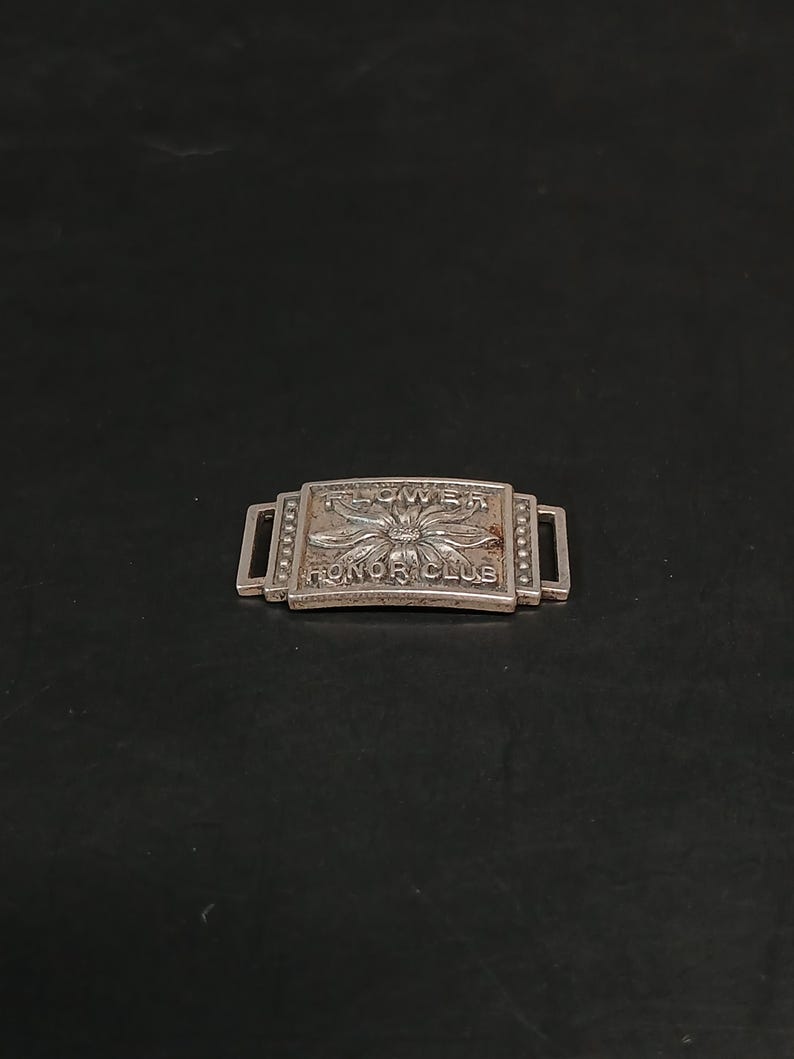 May include: A silver-toned metal bar with the words "FLOWER HONOR CLUB" engraved on it. The bar has a rectangular shape with a flower design in the center and two rectangular loops on each end.