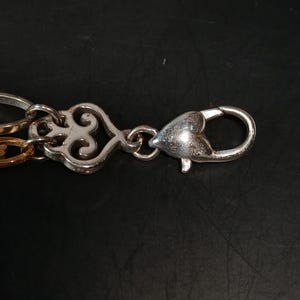 Puede incluir: Primer plano de un cierre y una cadena de metal plateado. El cierre tiene forma de coraz&oacute;n con una apertura de resorte. La cadena presenta eslabones decorativos ornamentados en tonos plateados y dorados. El fondo es de color oscuro y s&oacute;lido.