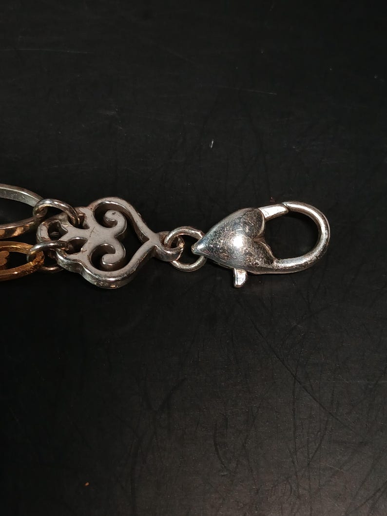 Puede incluir: Primer plano de un cierre y una cadena de metal plateado. El cierre tiene forma de coraz&oacute;n con una apertura de resorte. La cadena presenta eslabones decorativos ornamentados en tonos plateados y dorados. El fondo es de color oscuro y s&oacute;lido.