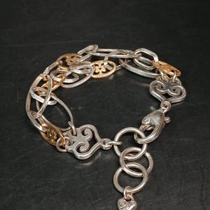 Puede incluir: Pulsera plateada con detalles dorados. La pulsera presenta una mezcla de eslabones ovalados y decorativos, con un dije en forma de coraz&oacute;n colgando del cierre. El dise&ntilde;o incorpora adornos de volutas y un cierre de mosquet&oacute;n.