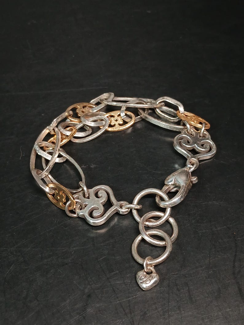 Puede incluir: Pulsera plateada con detalles dorados. La pulsera presenta una mezcla de eslabones ovalados y decorativos, con un dije en forma de coraz&oacute;n colgando del cierre. El dise&ntilde;o incorpora adornos de volutas y un cierre de mosquet&oacute;n.