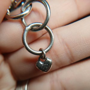 Puede incluir: Primer plano de una pulsera de cadena plateada con tres eslabones circulares entrelazados. Un peque&ntilde;o dije en forma de coraz&oacute;n cuelga del eslab&oacute;n inferior. La pulsera se sostiene en una mano, con el foco en la joyer&iacute;a.