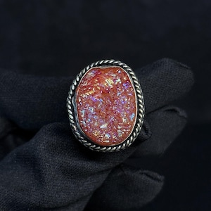 May include: An oval ring featuring a textured, iridescent orange druzy stone set within a twisted silver-toned metal border. The central stone displays a crystalline surface with shimmering highlights, framed by a decorative rope-style metal edge.