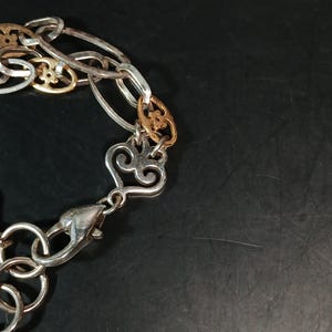 Puede incluir: Una pulsera plateada con una mezcla de eslabones ovalados y decorativos. Algunos eslabones son dorados con un dise&ntilde;o floral. La pulsera tiene un cierre en forma de coraz&oacute;n, sobre un fondo oscuro.