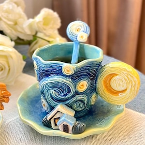 Puede incluir: Un juego de taza y platillo de cerámica con un diseño inspirado en "La noche estrellada" de Van Gogh. La taza, el platillo y la cuchara están pintados en tonos azules y amarillos, con patrones arremolinados y detalles de pequeñas casas. El asa es en espiral.
