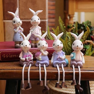 Könnte beinhalten: Sechs weiße Hasenfiguren mit langen Ohren, gekleidet in pastellfarbene Outfits. Einige Hasen sitzen mit baumelnden Beinen und halten Geschenke oder Bücher. Die Figuren sind auf einer Holzoberfläche und einem roten Buch angeordnet.