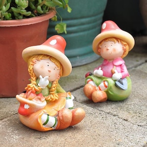 以下が含まれることがあります： キノコの帽子をかぶった子供たちの庭の置物2つ。オレンジ、黄色、緑、ピンクの色合いのカラフルな衣装を着ています。片方の置物は小さなシャベルを持ち、もう片方は顎に指を当てています。庭やパティオに最適です。
