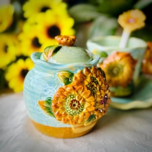 May include: A ceramic sugar bowl with a lid, painted in shades of blue, yellow, and green. The bowl is adorned with three-dimensional sunflowers. A matching cup and saucer are visible in the background, along with yellow sunflowers.