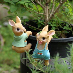 May include: Two rabbit figurines, one in a green shirt and the other in blue dungarees, clinging to a black plant pot. The bunnies have tan fur, pink ears, and carrot details. The scene is set in a garden with green foliage.