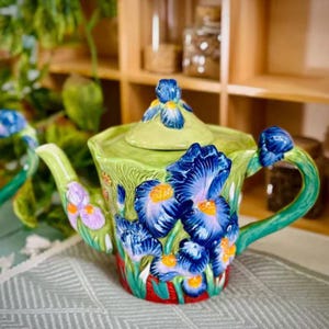 May include: A ceramic teapot featuring a green base and lid, decorated with blue iris flowers and orange centers. The handle and spout are green, and the base has red accents. The teapot has a floral design.
