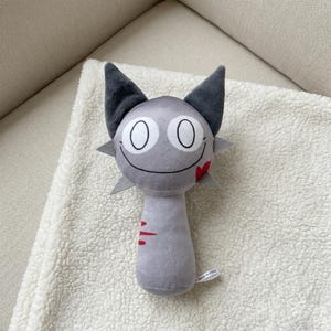 May include: A gray plush toy with a smiling face, large white eyes, and dark gray ears. The toy has a red heart-shaped detail and red slash marks. The toy is on a white, fluffy surface.