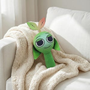 May include: A green plush toy shaped like a sprout with a smiling face, large eyes, and two leaf-shaped ears. The sprout has a stem and two leaves at the top, with a gradient of colors from green to orange. The toy is resting on a cream-colored knitted blanket.