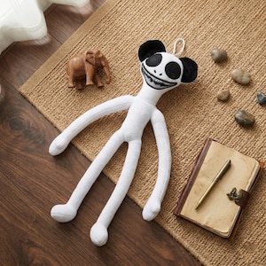 May include: A white plush toy with a black-and-white face and long limbs. The toy has black ears and a stitched, grinning mouth. It is lying on a brown woven rug, with a wooden elephant figurine and a journal nearby.