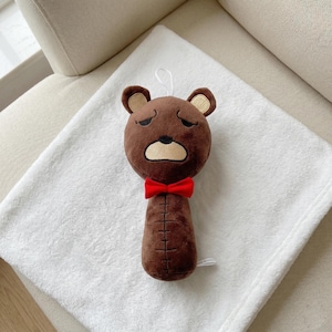 May include: A brown plush bear rattle with a red bow tie. The bear has a sad expression with dark brown eyes and a beige muzzle. The rattle is on a white blanket.