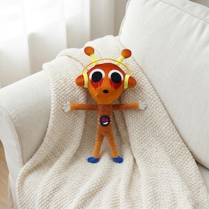 May include: Orange plush toy alien with large eyes, yellow headphones, and outstretched arms. The toy has small white hands, blue feet, and a red and black design on its chest. It is resting on a cream-colored blanket.