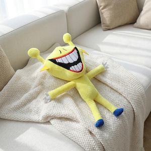 May include: A yellow plush toy with a large, open-mouthed smile, white teeth, and red gums. The toy has outstretched arms, small blue feet, and white hands. It is lying on a cream-colored blanket.