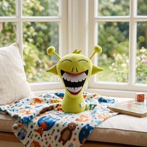 Op de afbeelding: Een limoengroen pluche speelgoed met een wijd open mond en grote witte tanden. Het speelgoed heeft twee antennes en ligt op een deken met een kleurrijk bosdierenpatroon. Een kinderboek en houten blokken zijn ook zichtbaar.