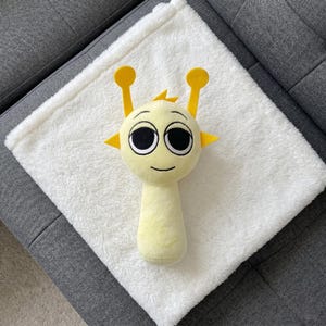 May include: A plush, yellow alien-like rattle toy with large black eyes, yellow antennae, and triangular ear accents, resting on a fluffy white blanket. The blanket is on a gray surface.