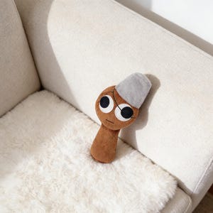 Puede incluir: Un peluche marrón con forma de seta con ojos grandes y redondos y una gorra gris. El juguete tiene una cara cosida y está sobre una superficie blanca texturizada.