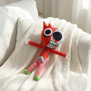 May include: A red plush doll with a half-skull face and open arms. The doll has a red body, pink legs, and green feet. The doll is resting on a white blanket.