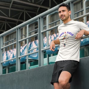 May include: A white t-shirt with the words "Track & Field" in blue and a graphic of a track athlete. The person is wearing black shorts and is leaning against a metal railing in a stadium setting.