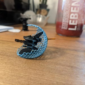 Op de afbeelding: Een zwart sciencefiction-voertuigmodel in miniatuur, omhuld door een lichtblauw, halfrond energieschild met een zeshoekig rasterpatroon. Het voertuig heeft een puntige voorkant, een kleine oranje antenne en staat op een zwarte ronde basis.