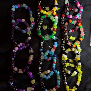 May include: A collection of colorful beaded bracelets with various text and star-shaped accents. The bracelets feature a mix of vibrant colors, including blue, purple, yellow, green, and red, with black and white letter beads spelling out names and phrases.