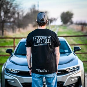 May include: Black t-shirt with a white license plate design that reads "TEXAS 1BAD+1LE THE LONE STAR STATE". The shirt is worn by a person standing in front of a silver car. The person is wearing blue jeans and red shoes.