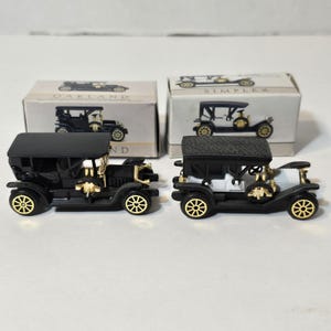 May include: Two miniature vintage car models. One is black with gold accents, the other is black and white with gold accents. Both have black tops and gold wheels. The boxes behind the cars have car illustrations.