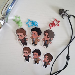May include: A collection of cartoon character stickers and star-shaped keychains in green, blue, and red. The stickers feature three different characters in brown, black, and tan coats. A black cord with a decorative pendant is also present.