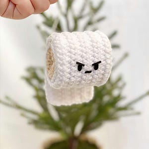 May include: A handmade, white crocheted toilet roll ornament with a black, angry face. The ornament is held up in front of a small, green Christmas tree. The toilet roll has a beige interior.