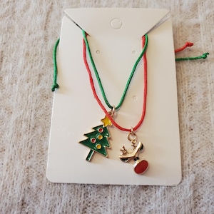 May include: Two Christmas-themed necklaces on a white card. One necklace features a green Christmas tree pendant with red and yellow accents. The other has a reindeer pendant with a red nose. Both necklaces have red and green cords.