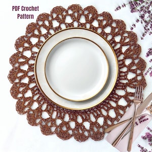May include: A brown, crocheted doily with a scalloped edge, beneath two white plates with gold rims. The text "PDF Crochet Pattern" is visible. A fork, knife, and spoon rest on a pink napkin, suggesting a dining setting.