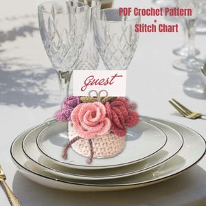 May include: A crochet flower arrangement place card holder featuring a light beige base with pink, mauve, and dark red roses. A white rectangular card with the red text "Guest" is held by a silver metal clip. Includes PDF Crochet Pattern + Stitch Chart.