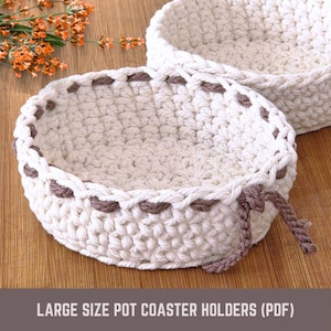 May include: Two off-white, crocheted pot coaster holders with brown trim and accents. The holders are round with a textured appearance and a braided edge. The text at the bottom reads "LARGE SIZE POT COASTER HOLDERS (PDF)".