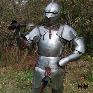 May include: A full suit of polished steel armor, including a helmet, chest plate, and leg protection. The armor is worn with chainmail and leather straps. A sword is held in the right hand. The background is a natural outdoor setting.