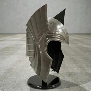 May include: A metallic silver helmet with a unique design, including layered, wing-like features and a pointed crest. The helmet is presented on a black circular stand, against a neutral backdrop. The helmet's surface reflects light, showcasing its detailed construction.