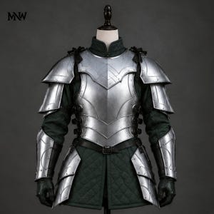 May include: A silver metal breastplate and shoulder armour set worn over a dark green quilted tunic. The armour features articulated pauldrons, a central chest plate, and matching arm guards with black leather straps and buckles. MNW logo is visible.