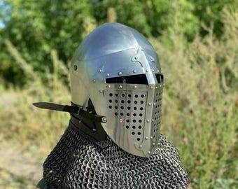 Puede incluir: Un casco medieval de acero pulido con visera remachada y cofia de cota de malla. El casco tiene una parte superior redondeada y una serie de pequeños remaches que aseguran la visera. Un pequeño cuchillo está sujeto al lateral. El fondo es follaje verde borroso.