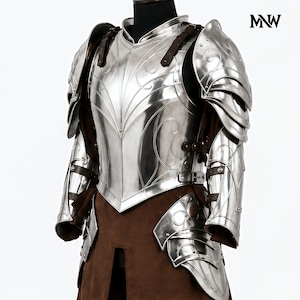 May include: A polished silver metal suit of armor featuring intricate engraved scrollwork patterns on the chest plate and shoulder pauldrons. The armor includes dark brown leather straps and is worn over a brown fabric tunic. The logo MNW is visible.