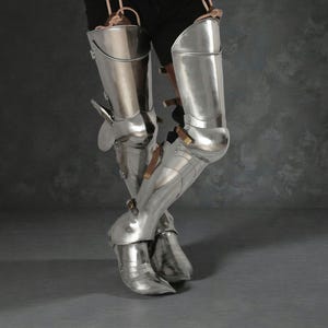 May include: Polished steel leg armor, featuring articulated plates for knee and lower leg protection. The armor includes leather straps and buckles for secure fastening. The design is reflective, with a medieval aesthetic.