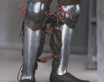 May include: Close-up of medieval leg armour. The armour is made of polished metal and brown leather. The knee and shin guards are connected with leather straps and red ties. The boots are brown leather.