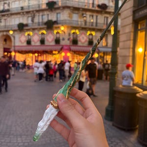 May include: A green and gold wand with a white handle. The wand has a twisted design with gold accents. The tip of the wand is a clear green color. The background is a blurred street scene with buildings and people.