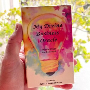 May include: A rectangular card deck box titled My Divine Business Oracle, A 60 Card Deck and Guidebook, created by Jane Yutrzenka Bruce. The box features a colorful watercolor lightbulb illustration in shades of pink, yellow, and blue.
