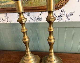 Puede incluir: Dos candelabros antiguos de latón. Cada candelabro tiene una base hexagonal, un tallo decorativo y una parte superior cilíndrica para una vela. Los candelabros se exhiben sobre una superficie de madera, con una pintura y papel tapiz en el fondo.