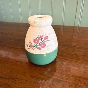May include: A small, decorative vase with a rounded body and a narrow opening. The top portion is off-white, while the bottom is painted green. Pink floral accents adorn the front, adding a touch of color and charm to this unique piece.