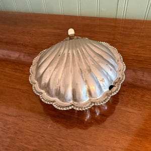 Op de afbeelding: Een zilverkleurige, schelpvormige schaal met een decoratieve, parelrand en een kleine, gebroken witte handgreep. De schaal heeft een geribbelde oppervlakte en staat op een houten oppervlak.