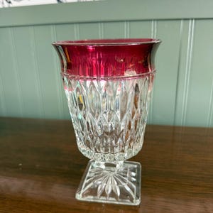May include: A clear glass vase with a ruby red rim. The vase has a square base and a textured, diamond-cut pattern on the body. The vase is likely vintage and suitable for displaying flowers or as a decorative piece.